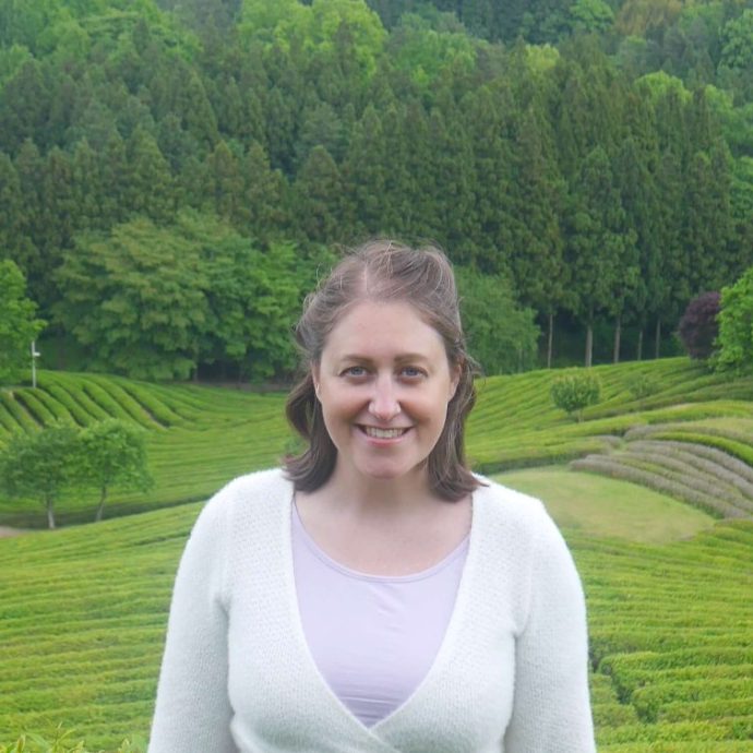 Meg smiles at the camera standing at the top of a slope of tea fields with a forest in the background