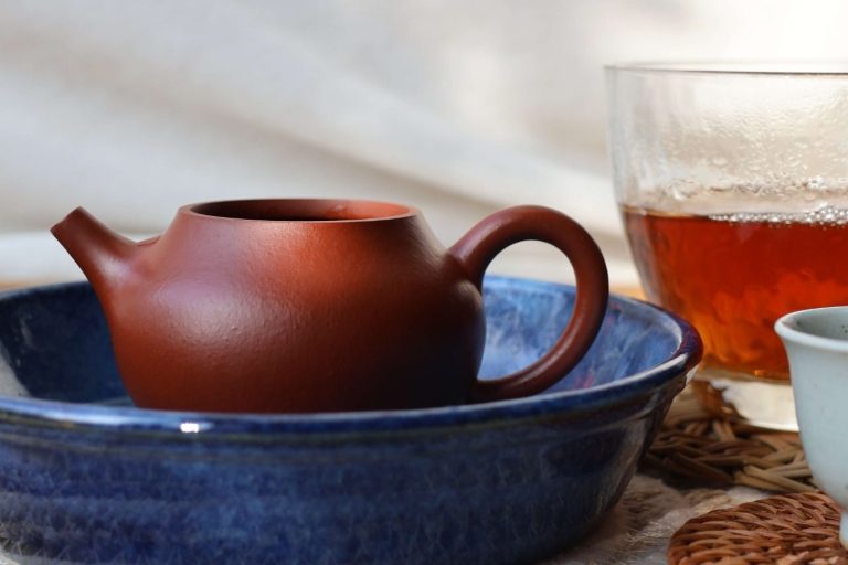 A red clay teapot in a blue ceramic tray, with a glass pitcher containing dark red-brown tea liquor and porcelain cup in the background