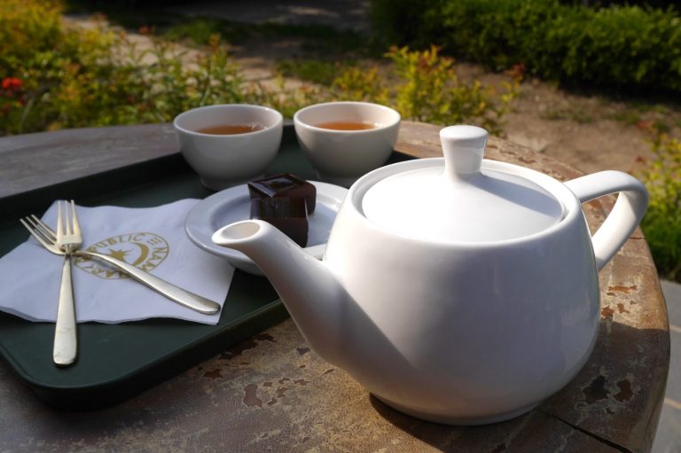 Close-up of a large Western-style white teapot, with two small cups and a plate with cake in the background