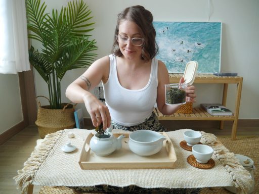 Meg is sat at a low table covered in a cloth, with a wooden tray holding teapot and pitcher. She adds loose leaf tea to the teapot.