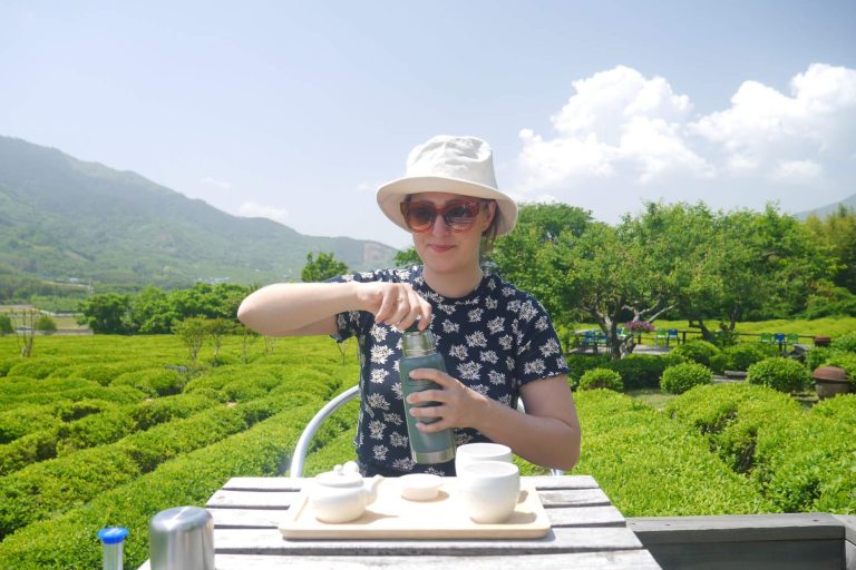 Meg sits at a small table with a tray containing a small teapot and cups, with tea fields in the background