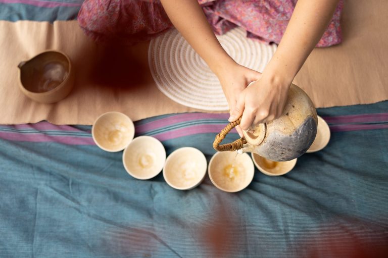 Overhead photo of Meg pouring hot water from a large stoneware kettle into a series of 6 bowls arrayed in a semicircle