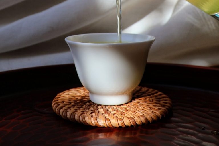 Tea being poured into a small white porcelain teacup on a bamboo coaster, with light falling across the cup and the rest of the table in shadow